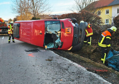 Einsatzkräfte arbeiten an einem umgestürzten roten Schulbus-Kleintransporter am Straßenrand.
