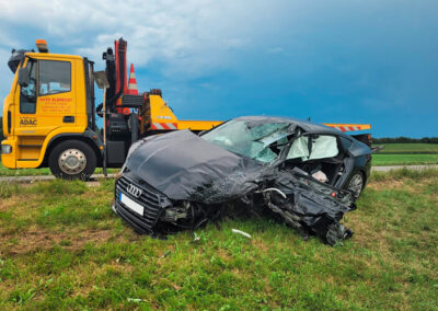 Schwer beschädigtes, dunkles Auto steht neben einem ADAC-Abschlepp-LKW auf einer Wiese.