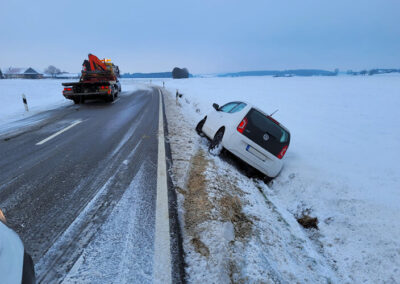 Ein weißer Kleinwagen steht nach einem Unfall in einem verschneiten Straßengraben, daneben ein ADAC-Abschlepp-LKW.