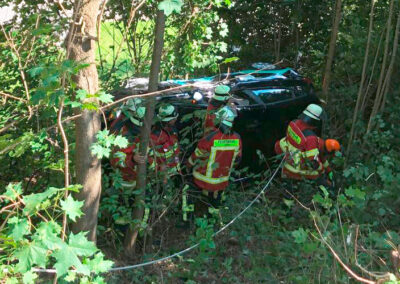 Dunkles Auto steht nach einem Unfall im Wald zwischen Bäumen, umringt von Einsatzkräften